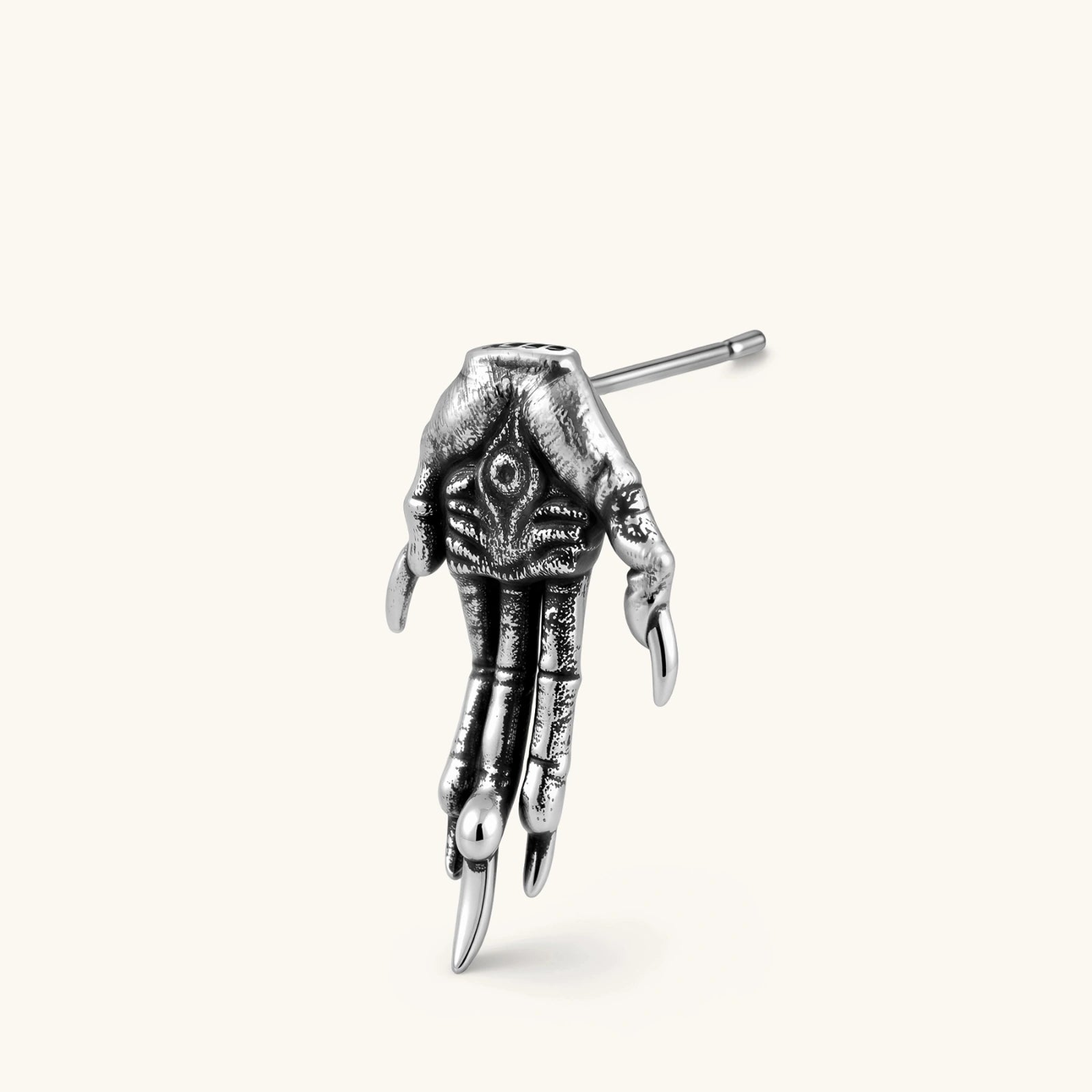 Silver stainless steel stud earring shaped like a skeletal demon claw hand with an eye motif on the palm and sharp talon fingers.