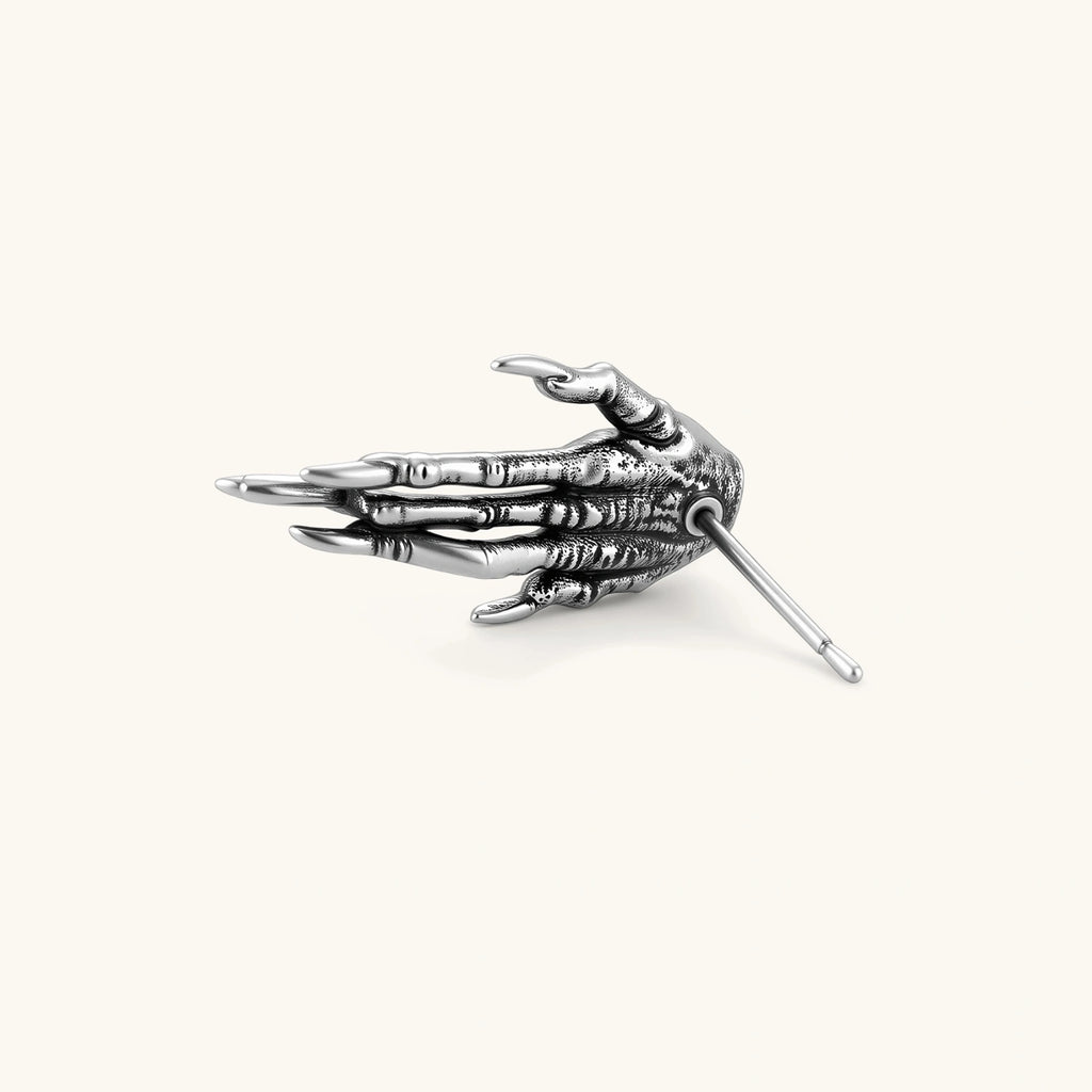 Silver stainless steel stud earring shaped like a skeletal demon claw hand with an eye motif on the palm and sharp talon fingers.
