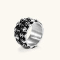 Wide stainless steel ring with raised 3D skull cluster design, black oxidized recesses, brushed silver interior, side angle view.