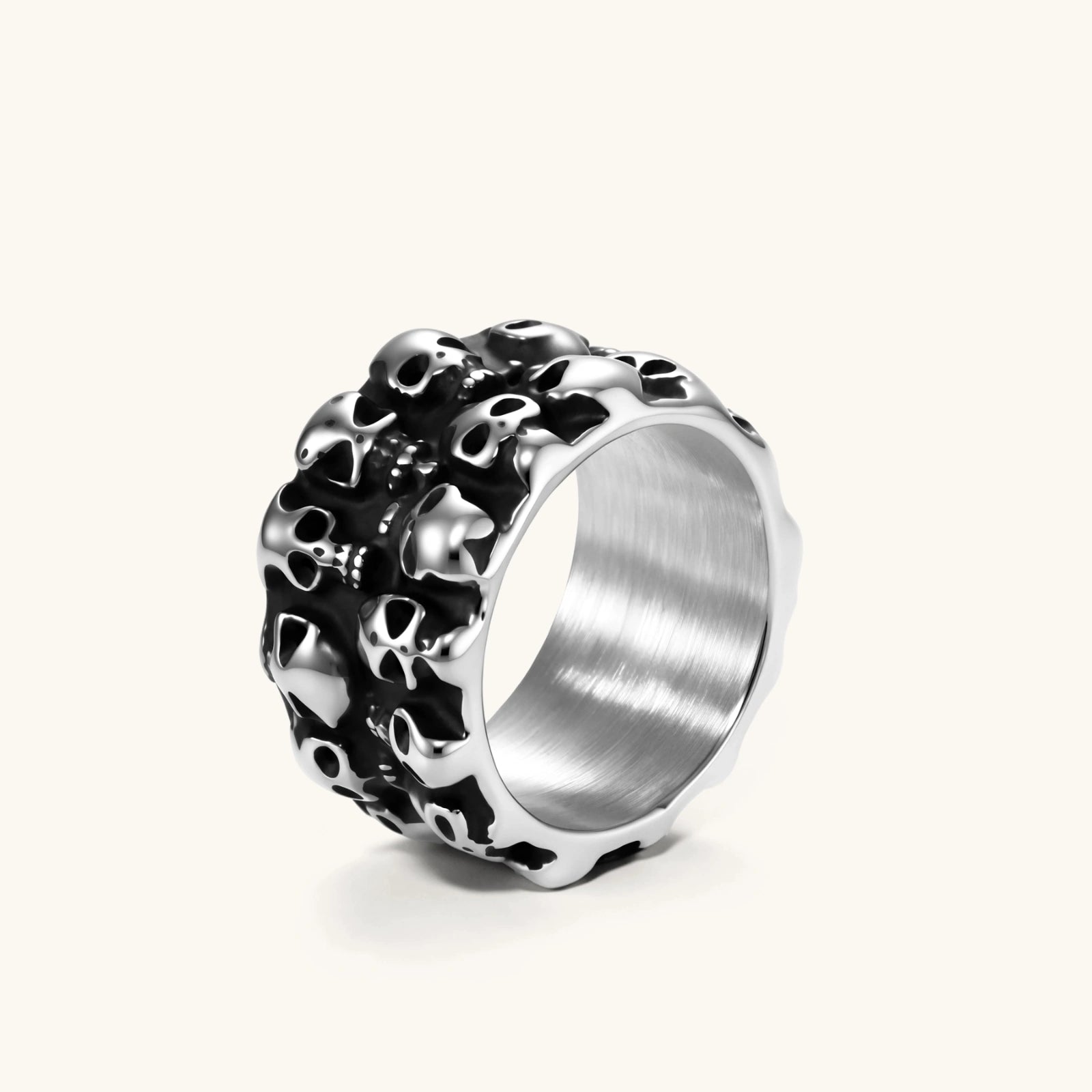 Wide stainless steel ring with raised 3D skull cluster design, black oxidized recesses, brushed silver interior, side angle view.